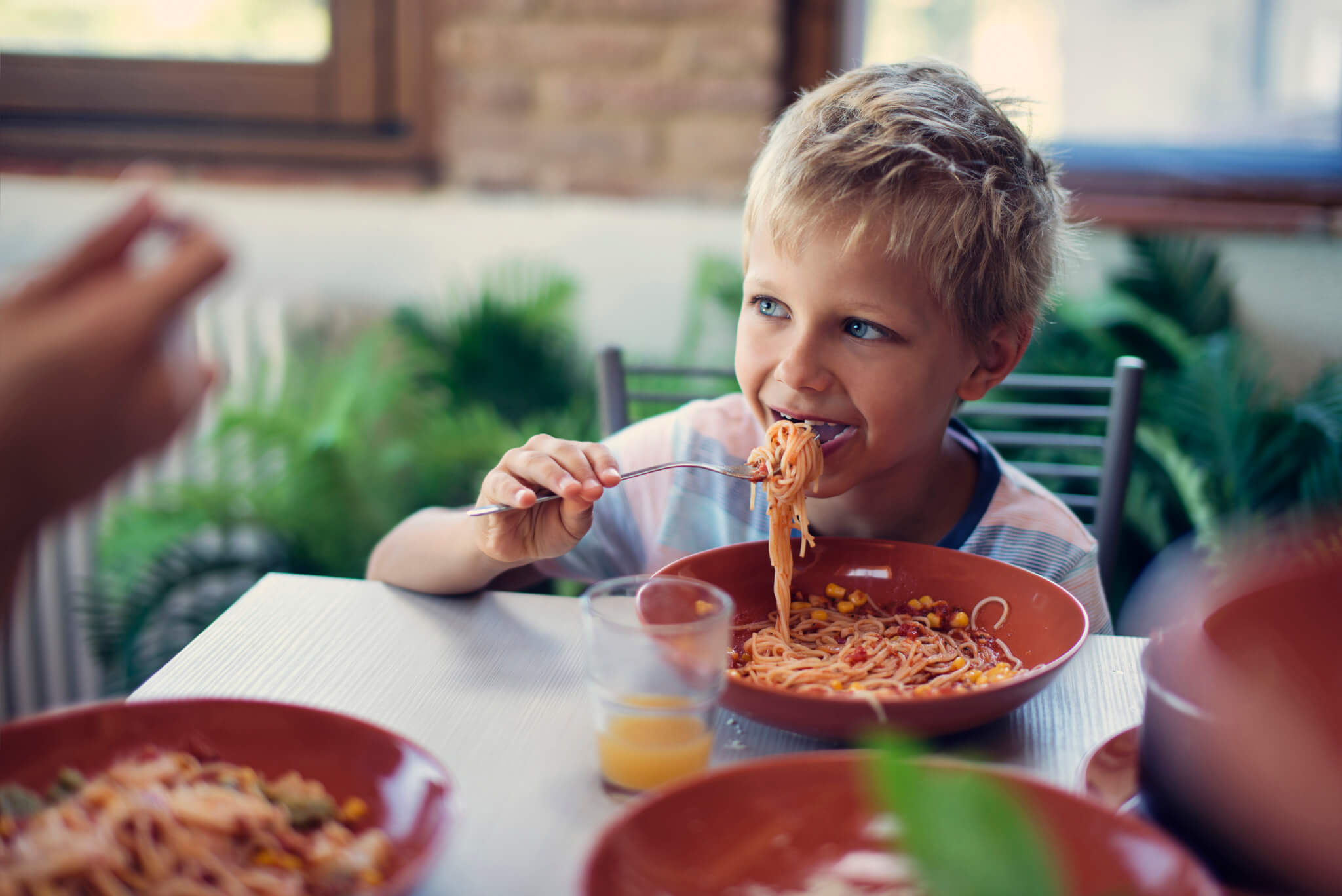 A young boy seated at the table using a fork to bring the spaghetti and tomato sauce to his mouth